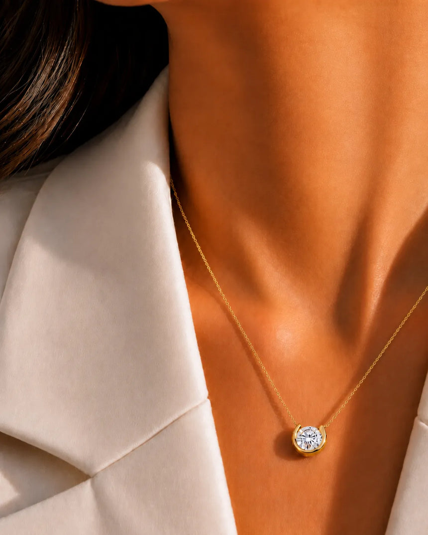 A close-up of a model's neck and collarbone wearing a delicate gold chain necklace with a round bezel-set clear gemstone pendant, styled with a cream-colored blazer. - SARATTI