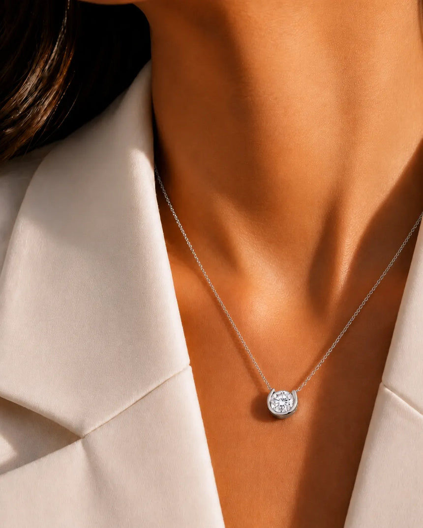 A close-up of a model's neck and collarbone wearing a delicate silver chain necklace with a round, bezel-set clear gemstone pendant, styled with a cream-colored blazer.