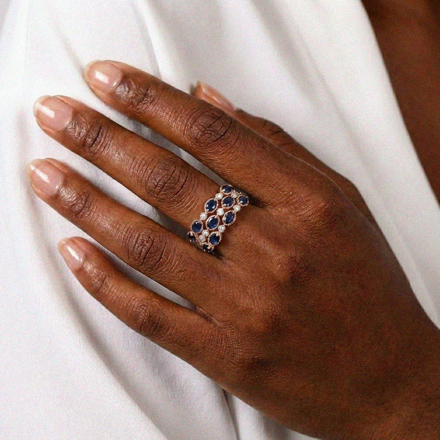 Hand wearing a ring with blue stones and clear stones on a white background - Multi row sapphire diamond ring rose gold finish | SARATTI