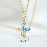 Gold necklace with a blue aquamarine gemstones designed into cross pendant on a light background, featuring the SARATTI brand.