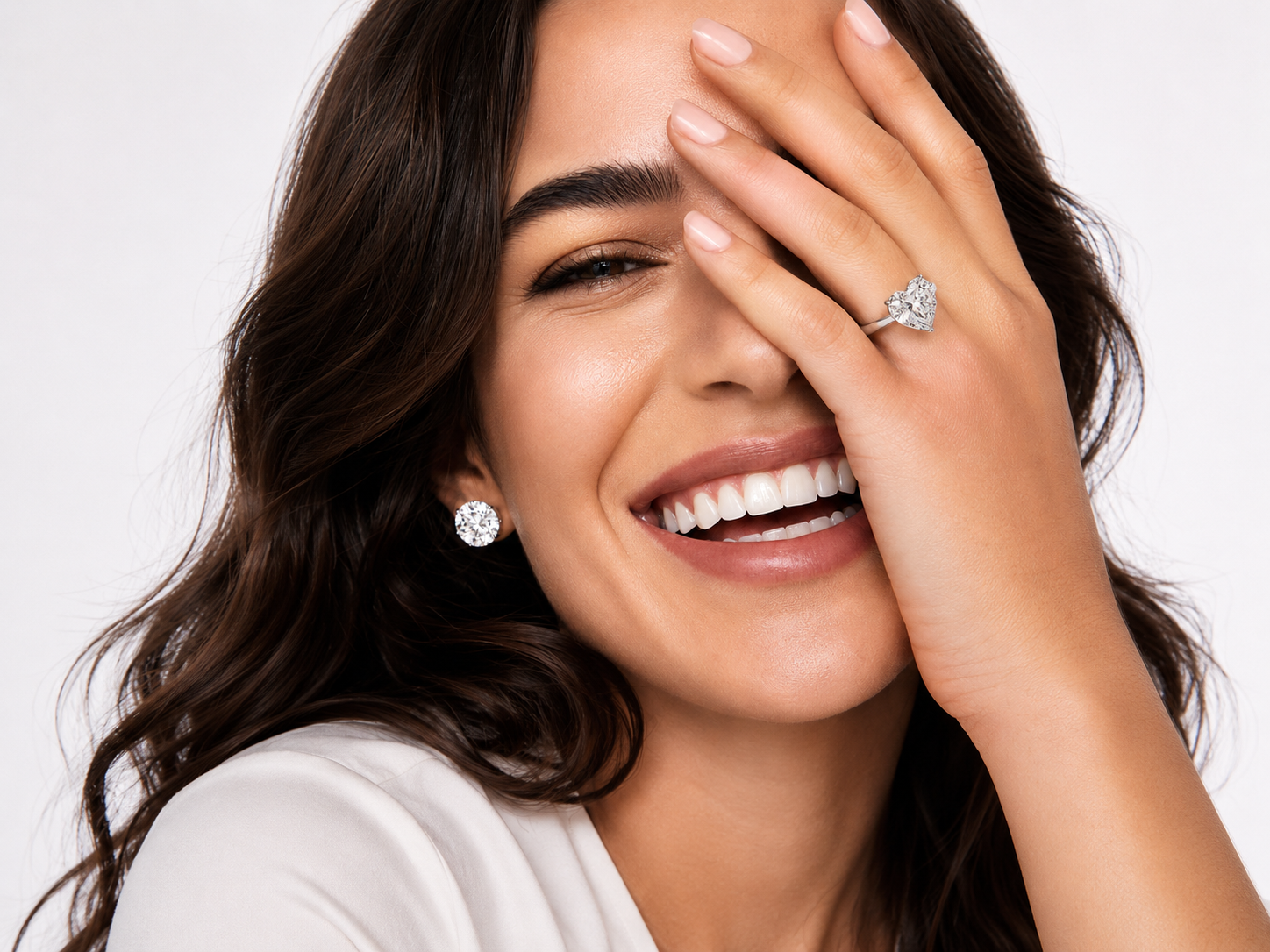 A close-up lifestyle shot of a woman with dark wavy hair, smiling broadly while covering one eye with her hand. She is wearing a sparkling diamond engagement ring on her ring finger and a matching round diamond stud earring. - SARATTI