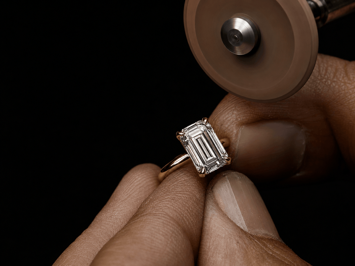 A close-up of a hand holding a yellow gold ring with an emerald-cut clear gemstone, while a rotary diamond cutting wheel approaches the setting against a black background. - SARATTI