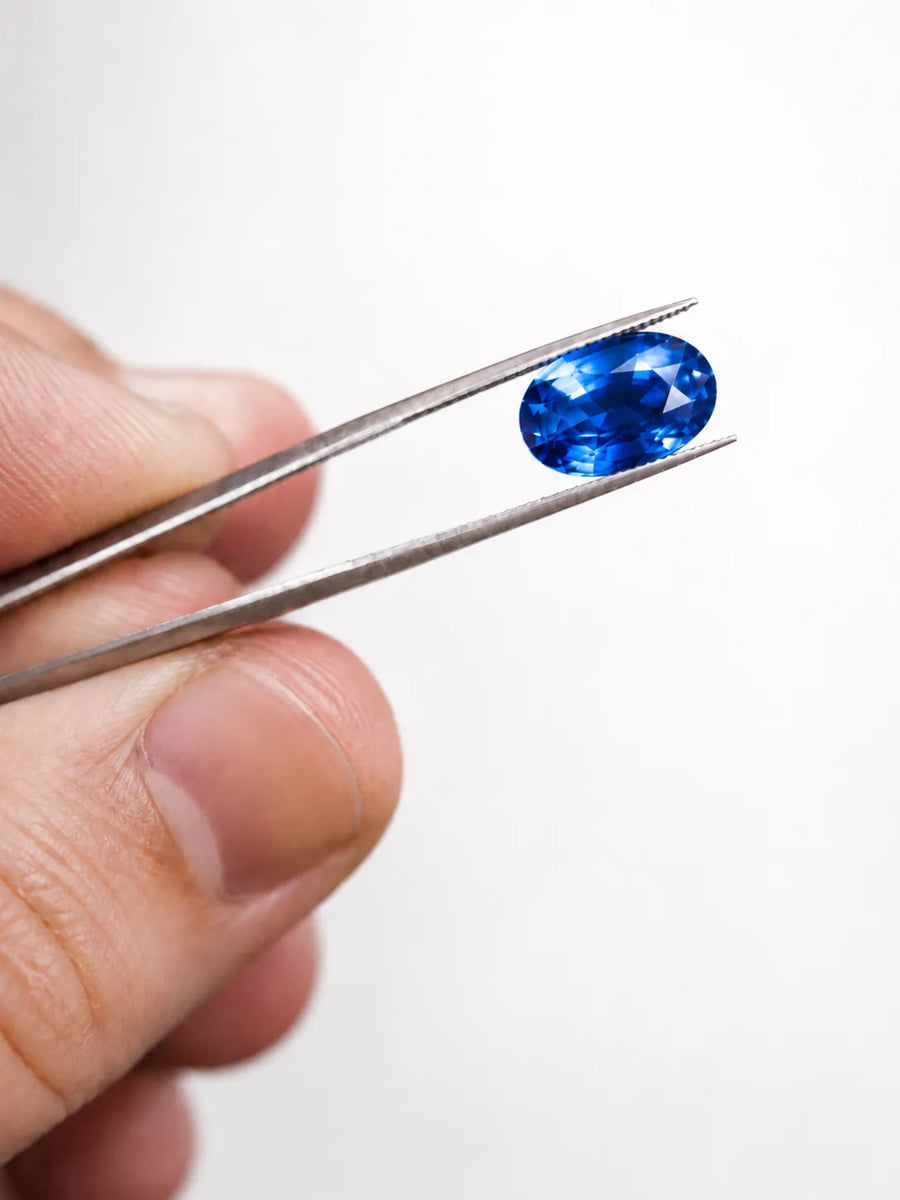 SARATTI hand-selected oval blue sapphire held in jeweler’s tweezers under natural daylight, showing honest color and clarity