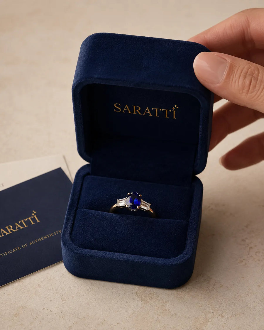 Oval blue sapphire and diamond SARATTI ring nestled inside a navy velvet presentation box beside certificate of authenticity card