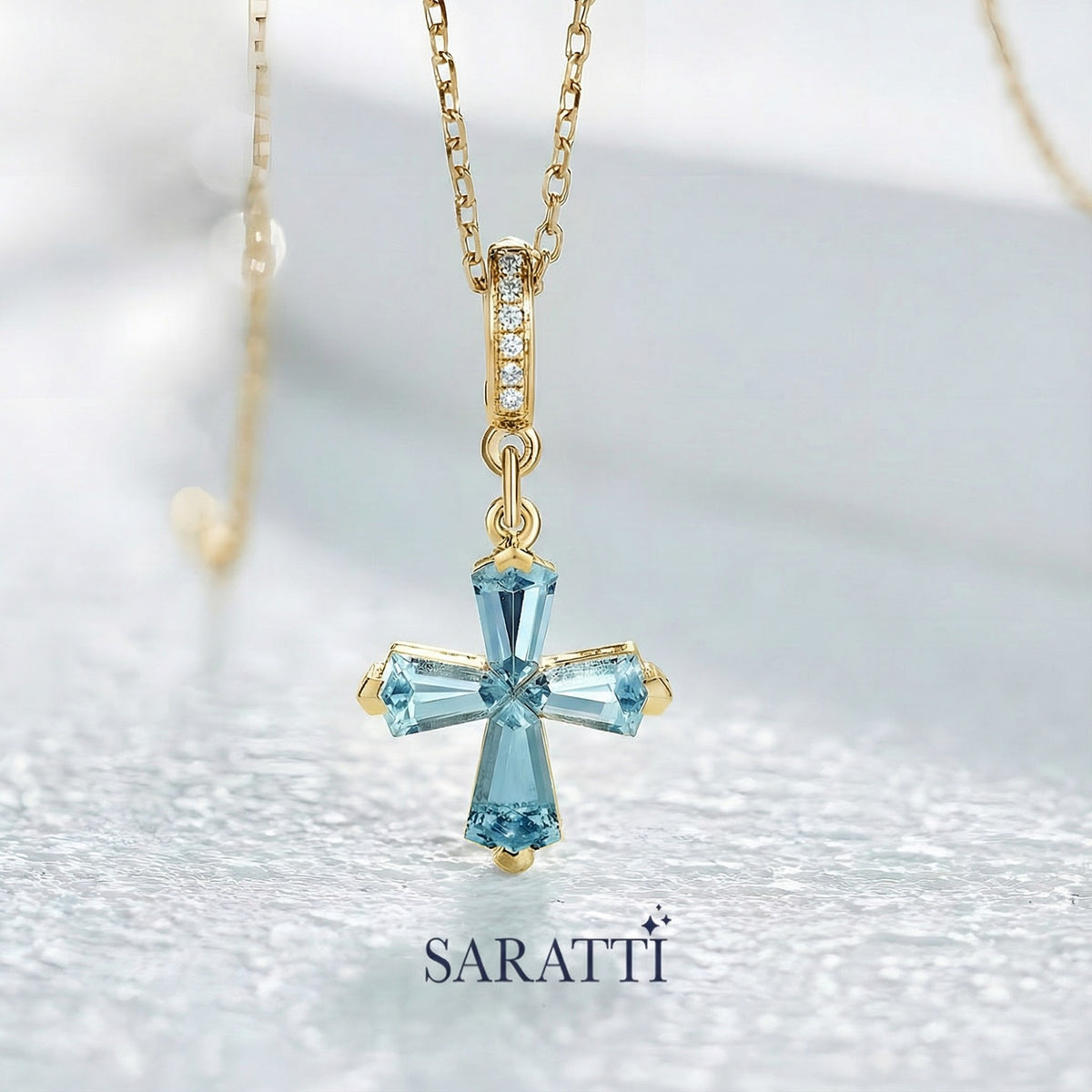 Gold necklace with custom-cut blue aquamarine gemstones designed into a cross pendant on a soft white background, featuring the brand 'Saratti'.